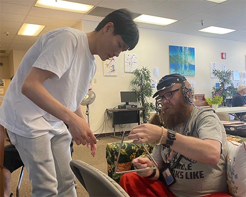 A community member with a disability learns to play the triangle with the help of a student
